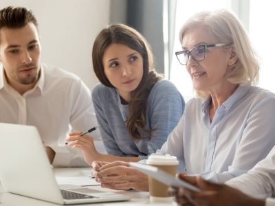 Focused young students interns workers making notes listening to old senior aged female manager coach mentor leader teacher talking at group office meeting instructing business work team with laptop
